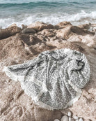 Die Decke Savanna von Main Design, eine grau-weiß gemusterte Decke mit Fransen, liegt auf felsigem Sand in der Nähe des Meeres, während im Hintergrund die Wellen rauschen.