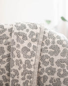 Eine Nahaufnahme der gefalteten, hellen Decke Savanna von Main Design mit grauem Leopardenmuster, drapiert auf einer Fläche mit sanft verschwommenem Hintergrund.