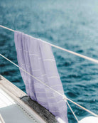 Ein Maxi Strandtuch De La Mer von Main Design mit lavendelfarbenen Streifen aus hochwertiger Baumwolle hängt über dem Bootsreling, im Hintergrund ruhiges blaues Wasser.