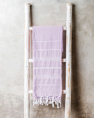Ein hellviolettes Maxi Strandtuch De La Mer von Main Design aus hochwertiger Baumwolle mit weißen Streifen und Fransen liegt über einer weißen Holzleiter vor einer grauen Wand.