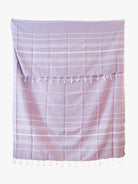 Das Maxi Strandtuch De La Mer von Main Design ist ein lavendelfarbenes Strandtuch aus 100% Baumwolle mit dünnen weißen Streifen und Quasten am oberen und unteren Rand. Es ist OEKO-TEX-zertifiziert und liegt flach auf einem weißen Hintergrund.