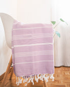 Das Maxi Strandtuch De La Mer von Main Design aus 100% Baumwolle mit weißen Fransen, OEKO-TEX zertifiziert, ist über einen weißen Stuhl auf Parkett drapiert. Im Hintergrund sind eine grüne Pflanze und ein weißer Vorhang sichtbar.