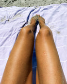 Auf dem Main Design Maxi Strandtuch De La Mer, aus 100% Baumwolle mit dünnen weißen Streifen, OEKO-TEX zertifiziert, werden die sandigen Beine einer Person am Strand mit Sand im Hintergrund ausgestreckt.