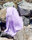 Ein großer Felsen ist mit dem hellvioletten Maxi Strandtuch De La Mer von Main Design zwischen grauen Felsen drapiert, mit trockenem Gras und Sträuchern im Hintergrund.
