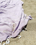 Das lilafarbene Maxi Strandtuch De La Mer von Main Design aus 100% Baumwolle mit weißen Streifen und geknoteten Fransen liegt auf dem Sand, etwas Sand bedeckt das OEKO-TEX-zertifizierte Strandtuch.