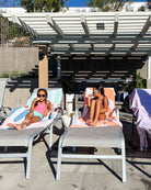 Zwei junge Mädchen in Badeanzügen entspannen sich am Pool und genießen ein Eis auf Liegestühlen, die mit Maxi Strandtuch De La Mer von Main Design bezogen sind. Bunte Handtücher und helles Sonnenlicht sorgen für einen verspielten Touch unter der Pergola.