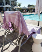 Ein Loungesessel am Pool mit einem lila-weiß gestreiften Maxi Strandtuch De La Mer von Main Design aus 100 % Baumwolle und mit OEKO-TEX-Zertifikat ist auf einer sonnigen Terrasse in der Nähe des Pools drapiert, mit weiteren Stühlen und Sonnenschirmen im Hintergrund.