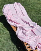 Das OEKO-TEX zertifizierte Maxi Strandtuch De La Mer von Main Design aus 100% Baumwolle in Pink-Weiß mit Tasseln liegt über einem Holz-Loungesessel auf grünem Rasen im Sonnenlicht.