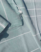 Eine Nahaufnahme des Maxi Strandtuchs De La Mer von Main Design aus hellblau-grüner OEKO-TEX-Baumwolle mit weißen Streifen, ausgefransten Rändern und sichtbarer Textur, teilweise gefaltet und im Sonnenlicht.