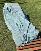Ein Loungesessel im Gras ist mit dem hellblau gestreiften Maxi Strandtuch De La Mer von Main Design drapiert, das an den Enden gerafft ist und den Sessel vollständig bedeckt, während das Sonnenlicht in der Nähe Schatten wirft.