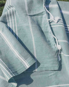 Eine Nahaufnahme des hellgrün-weiß gestreiften Maxi Strandtuchs De La Mer von Main Design aus OEKO-TEX-Baumwolle mit Quasten, drapiert im Freien im Sonnenlicht, wobei die schnell trocknende Textur hervorgehoben wird.