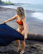 Eine Frau breitet den Main Design Sarong Bali an einem Sandstrand in der Nähe des Wassers aus. Sie trägt eine Sonnenbrille, ein orangefarbenes Bikinioberteil und eine karierte Hose an einem bewölkten Tag.