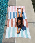 Ein Mädchen lächelt in die Kamera, während es auf dem Bauch am Pool des Main Design Strandtuch Blockberry liegt. Die pastellblauen, rosa, weißen und braunen Streifen liegen auf dem Beton neben dem klaren blauen Wasser.