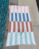 Das Strandtuch Blockberry von Main Design mit pastellfarbenen rosa, blauen, braunen und grünen Streifen liegt flach auf Beton neben einem Swimmingpool mit klarem blauen Wasser.