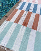 Ein Main Design Strandtuch Blockberry mit blauen, weißen, rostfarbenen und hellgrünen Streifen liegt auf dem Beton des Pools neben dem glitzernden Wasser.