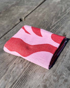 Ein gefaltetes Strandtuch Coral Flow von Main Design mit rosafarbenem und rotem Muster aus 100 % Baumwolle ruht auf einer verwitterten Holzoberfläche im hellen Sonnenlicht.