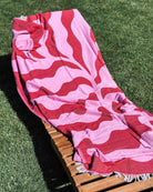 Ein hölzerner Loungesessel im Gras, über den das Main Design Strandtuch Coral Flow mit seinen rosa und roten Wellenmustern drapiert ist und das Sonnenlicht einfängt.