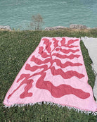 Ein Strandtuch der Serie „Main Design“ mit dem Motiv „Coral Flow“, rosa Jacquard-Einfassung und wellenförmigen roten Mustern liegt auf grünem Gras in der Nähe eines hellblauen Gewässers und einiger Felsen; rechts ist teilweise ein weiteres gestreiftes Handtuch zu sehen.