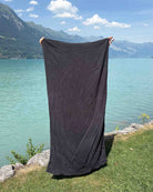 Eine Person hält ein großes „Main Design Strandtuch Dune“-Handtuch hoch und steht dabei auf einer Wiese an einem türkisfarbenen See, hinter ihr sind Berge und der blaue Himmel zu sehen. Über dem Handtuch sind nur ihre Hände zu sehen.