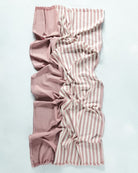 Ein zerknittertes rechteckiges Strandtuch Fiesta von Main Design, halb einfarbig staubrosa, halb rosa und weiß gestreift, auf einer hellen Oberfläche. Hergestellt aus Baumwolle und OEKO-TEX-zertifiziert für Qualität und Sicherheit.