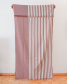 Das Main Design Strandtuch Fiesta aus OEKO-TEX-Baumwolle zeigt vertikale cremefarbene und gedeckte rosa Streifen auf der einen Seite und einfarbiges gedecktes Rosa auf der anderen Seite, die vor einer weißen Wand und einem Holzboden hängen.