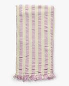Das Strandtuch Melody von Main Design ist ein gefaltetes, rechteckiges Hamamtuch mit vertikalen hellvioletten und cremefarbenen Streifen aus recycelter Baumwolle und kurzen Fransen am Ende.
