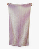 Das Strandtuch Melody von Main Design aus recycelter Baumwolle mit lavendel-weißen Streifen, kleinen Fransen und leichter Knitteroptik hängt vor schlicht weißem Hintergrund.