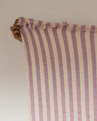 Eine Hand hält das Main Design Strandtuch Melody hoch, ein Strandtuch aus recycelter Baumwolle mit vertikalen lavendelfarbenen und cremefarbenen Streifen und ausgefransten Rändern vor einem schlichten hellen Hintergrund.