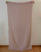 Ein Main Design Strandtuch Melody in beigen und lavendelfarbenen Längsstreifen aus recycelter Baumwolle steht an einer hellen Wand mit ausgefransten Kanten und einem Holzboden darunter.
