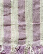 Eine Nahaufnahme des Strandtuchs Melody von Main Design zeigt abwechselnd hellviolette und cremefarbene Streifen mit einer Mischung aus lila und weißen Fransen, die aus recycelter Baumwolle gefertigt sind.