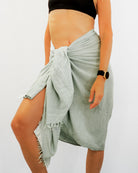 Eine Person in einem schwarzen bauchfreien Oberteil und dem hellgrünen Strandtuch Nomad Sarong von Main Design, der in der Taille gebunden ist, trägt eine Uhr am linken Handgelenk. Dieses stilvolle Ensemble aus OEKO-TEX Baumwolle bietet Komfort und ist der ideale Reisebegleiter für jedes Abenteuer.