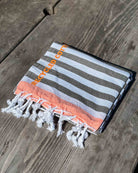 Das Strandtuch Ocean Loop von Main Design aus recycelter Baumwolle zeigt weiße, graue und orangefarbene Streifen mit ausgefransten Quasten und dem Text RECYCLED COTTON" auf einer Holzoberfläche.