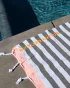 Ein Strandtuch „Ocean Loop“ von Main Design mit Streifenmuster und dem Label „RECYCLED COTTON“ liegt am Rand eines Pools – teilweise im Sonnenlicht, während im Hintergrund das blaue Wasser und die Poolfliesen zu sehen sind.