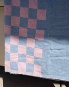 Ein „Strandtuch Pixel“ von Main Design mit einem modernen Schachbrettmuster in Rosa und Blau hängt so, dass ein Teil des Handtuchs im Sonnenlicht und ein Teil im Schatten liegt, wodurch seine Struktur und der auffällige Farbkontrast besonders gut zur Geltung kommen.