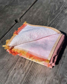 Ein gefaltetes rosafarbenes Strandtuch Riviera von Main Design mit gelben und orangefarbenen Fransen ruht auf Holz im natürlichen Sonnenlicht. Aus 100% Baumwolle gefertigt, bringt dieses Handtuch einen lebendigen Stil in Ihre sonnigen Tage im Freien.