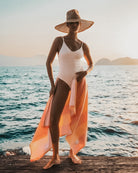 Eine Person in weißem Badeanzug und Strohhut steht bei Sonnenuntergang auf einem Steg am Meer und hält das Main Design Strandtuch Riviera - ein orange und rosa gestreiftes, schnell trocknendes Handtuch aus 100 % Baumwolle - in der Hand, mit den Bergen im Hintergrund.