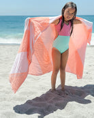 Ein junges Mädchen steht an einem sonnigen Strand in der Nähe des Meeres, trägt einen rosafarbenen und mintgrünen Badeanzug und hält das Main Design "Strandtuch Serra" - ein leichtes, kompaktes Strandtuch aus recycelter Baumwolle - offen hinter sich.