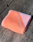 Das Strandtuch Serra von Main Design, ein gefaltetes pink-orangefarbenes Strandtuch aus recycelter Baumwolle mit teilweise sichtbarem "recycle cotton", sitzt auf einer Holzunterlage in hellem Sonnenlicht-leicht und kompakt für den einfachen Transport.