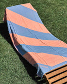 Ein Loungesessel mit dem Main Design Strandtuch Serra, einem leichten und kompakten Strandtuch mit kräftigen rosa und blauen Diagonalstreifen, steht auf grünem Gras, das von der Sonne beschienen wird, und einem kleinen Holzteil am Fußende.