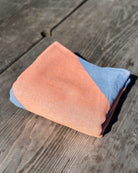 Ein gefaltetes Strandtuch Serra von Main Design aus recycelter Baumwolle mit pfirsichfarbenen und blauen Abschnitten liegt auf einer verwitterten Holzfläche im hellen Sonnenlicht und wirkt leicht und kompakt.