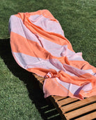 Ein hölzerner Loungesessel auf grünem Gras ist locker mit dem Strandtuch Serra von Main Design drapiert, einem orange-weiß gestreiften Handtuch, das sich teilweise im Sonnenlicht faltet.
