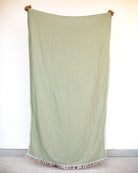 Eine Person steht vor einer weißen Wand und hält das große, hellgrüne Strandtuch Zen von Main Design hoch. Das Handtuch aus OEKO-TEX-Baumwolle hat ausgefranste Ränder, hinter dem strandtauglichen Stoff sind nur ihre Hände und Schuhspitzen zu sehen.