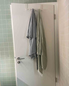 An einer weißen Tür mit zwei Haken hängen ein gestreiftes Hemd und ein hellgrünes Main Design Strandtuch Zen-Handtuch. Die Tür befindet sich in einem Badezimmer mit grün und beige gefliesten Wänden.