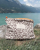 Eine „Aloha Collection“-Tasche „Aloha Mid Pouch“ mit Leopardenmuster liegt im Gras neben einem türkisfarbenen See, umrahmt von Bergen und einem teilweise bewölkten Himmel.