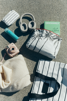 Flatlay-Aufnahme von Reiseutensilien auf einer gesprenkelten Oberfläche, darunter eine gestreifte Kosmetiktasche, die „Aloha Mid Pouch“ aus der Aloha-Kollektion, Kopfhörer, ein grünes Buch, eine türkisfarbene Wasserflasche, eine rosa Sofortbildkamera, ein cremefarbener Pullover und eine gestreifte Tragetasche.