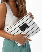 Eine Person, die ein weißes, ärmelloses Oberteil und eine blaugrüne Hose trägt, hält vor einem schlichten weißen Hintergrund eine „Aloha Collection Aloha Mid Pouch“ in die Hand, die weiße und schwarze Streifen sowie ein schwarzes Etikett aufweist.