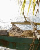 Eine „Aloha Mid Pouch“ aus der Aloha-Kollektion mit einem lebhaften tropischen Muster liegt am Rand eines alten Holzbootes am Wasser – ideal als Kosmetiktasche oder Reise-Organizer. Über ihr wiegen sich Palmblätter im Wind, während das Sonnenlicht auf dem Meer glitzert und einen idyllischen Inselmoment einfängt.