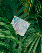 Ein weißes Kissen mit einem blau-lila Monstera-Blatt im Aquarellstil ruht auf grünem tropischem Blattwerk und wird durch die „Aloha Mid Pouch“ aus der Aloha-Kollektion ergänzt – für stilvolles Reiseflair inmitten üppiger Blätter.