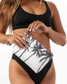 Eine Frau in einem schwarzen Bikini hält eine „Aloha Collection Aloha Small Pouch“ in der Hand, eine kleine Strandtasche mit Palmenmotiv. Ihre Fingernägel sind weiß lackiert, und an einem Handgelenk trägt sie Armbänder. Der Hintergrund ist weiß.