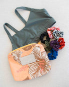 Ein grüner Sport-BH, die „Aloha Collection Aloha Small Pouch“ mit einem pfirsichfarbenen Blumenmuster und ein Stapel bunter Haargummis sind auf einer leicht strukturierten Oberfläche angeordnet. Diese stilvolle „Aloha Small Pouch“ ist zudem eine praktische, wasserfeste Tasche, die sich ideal als Bikini-Tasche für deine wichtigsten Utensilien eignet.